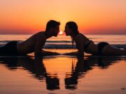 Perché in vacanza ci si innamora più facilmente (e come capire se è reciproco) Coppia sdraiata sulla spiaggia al tramonto, sguardi speculari che comunicano un'attrazione reciproca durante le vacanze estive