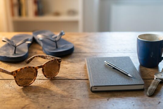 Sindrome da rientro: la statistica del 35% che gira da anni è una bufala Occhiali da sole con sabbia e infradito accanto ad agenda, penna, tazza di caffè e orologio su scrivania in legno, simbolo del passaggio dalle vacanze al rientro