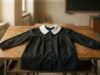 Grembiule nero delle medie: il vero motivo (non ideologico) per cui era quel colore Grembiule nero da scuola media con colletto bianco disteso su un banco di legno d'epoca in un'aula scolastica