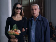 “La sera a Roma”: Vanzina cambia città e decennio al suo giallo dopo “Sotto il vestito niente” Scena del film "La sera a Roma" di Enrico Vanzina: una donna con occhiali da sole e fiori accanto a un uomo in giacca blu