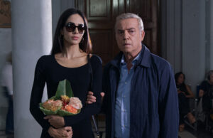“La sera a Roma”: Vanzina cambia città e decennio al suo giallo dopo “Sotto il vestito niente” Scena del film "La sera a Roma" di Enrico Vanzina: una donna con occhiali da sole e fiori accanto a un uomo in giacca blu