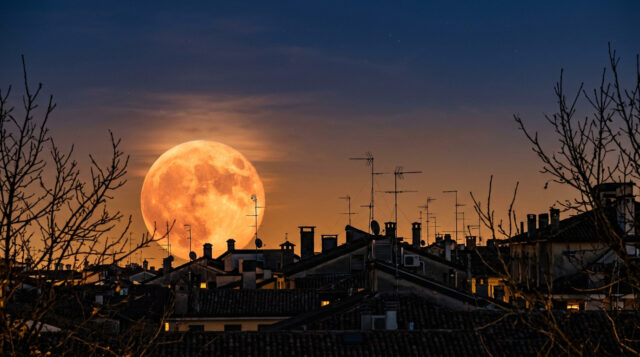 Luna del Cacciatore gigante e arancione che sorge dietro i tetti di un centro storico italiano al tramonto