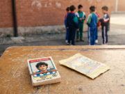 Il mercato dei doppioni delle figurine Panini: il vero rito che univa i cortili di scuola Mazzo di figurine calcistiche vintage anni '90 legato con un elastico su un vecchio banco di scuola, con una bustina vuota aperta a fianco e un gruppo di ragazzini in tuta sportiva sullo sfondo contro un muro di mattoni