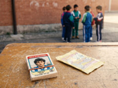 Il mercato dei doppioni delle figurine Panini: il vero rito che univa i cortili di scuola Mazzo di figurine calcistiche vintage anni '90 legato con un elastico su un vecchio banco di scuola, con una bustina vuota aperta a fianco e un gruppo di ragazzini in tuta sportiva sullo sfondo contro un muro di mattoni