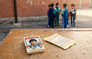 Il mercato dei doppioni delle figurine Panini: il vero rito che univa i cortili di scuola Mazzo di figurine calcistiche vintage anni '90 legato con un elastico su un vecchio banco di scuola, con una bustina vuota aperta a fianco e un gruppo di ragazzini in tuta sportiva sullo sfondo contro un muro di mattoni