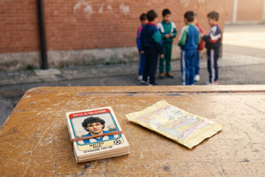 Il mercato dei doppioni delle figurine Panini: il vero rito che univa i cortili di scuola Mazzo di figurine calcistiche vintage anni '90 legato con un elastico su un vecchio banco di scuola, con una bustina vuota aperta a fianco e un gruppo di ragazzini in tuta sportiva sullo sfondo contro un muro di mattoni