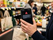 Sms falso che sembra dell’Agenzia delle Entrate: il dettaglio nel testo che lo tradisce Smartphone con falso avviso di allarme urgente in mano durante la spesa, esempio di truffa sms