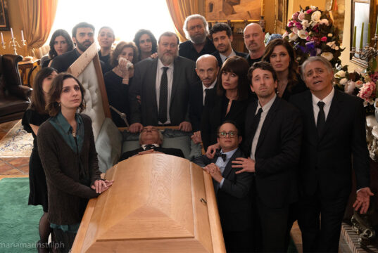 La nuova commedia di Massimiliano Bruno è al cinema da oggi, ma la storia non è affatto nuova Il cast di Un tranquillo funerale di famiglia riunito attorno alla bara durante una scena del film di Massimiliano Bruno