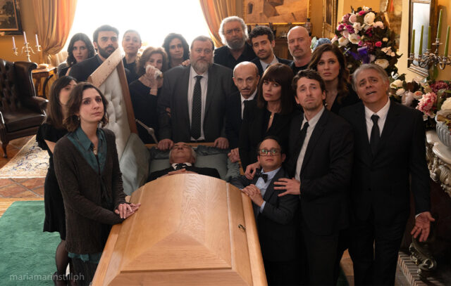 Il cast corale di Un tranquillo funerale di famiglia in una scena del film Il cast di Un tranquillo funerale di famiglia riunito attorno alla bara durante una scena del film di Massimiliano Bruno