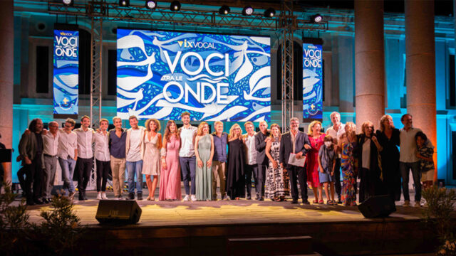 Gruppo di doppiatori e professionisti del cinema sul palco del festival "Voci tra le onde" Un gruppo di doppiatori, direttori di doppiaggio e professionisti del cinema in posa sul palco durante un'edizione passata del festival Voci tra le onde, con un grande schermo alle loro spalle che mostra il logo dell'evento